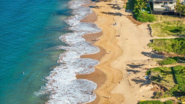 Une vue aérienne d’une plage avec une eau d’un bleu limpide, un rivage sablonneux et quelques personnes se promenant le long de la plage.