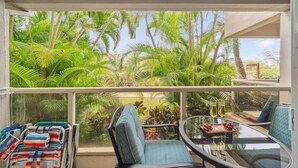Un balcon donnant sur une végétation tropicale luxuriante, une table en verre sur laquelle est disposée une corbeille de fruits et des chaises aux couleurs vives.