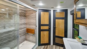 A modern bathroom with a glass shower enclosure, two wooden doors with ventilation slats, a bench, and a sink area with a mirror and towel.