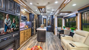 A modern RV interior with a kitchen, dining area, and living space, featuring wood flooring, built-in appliances, and large windows.