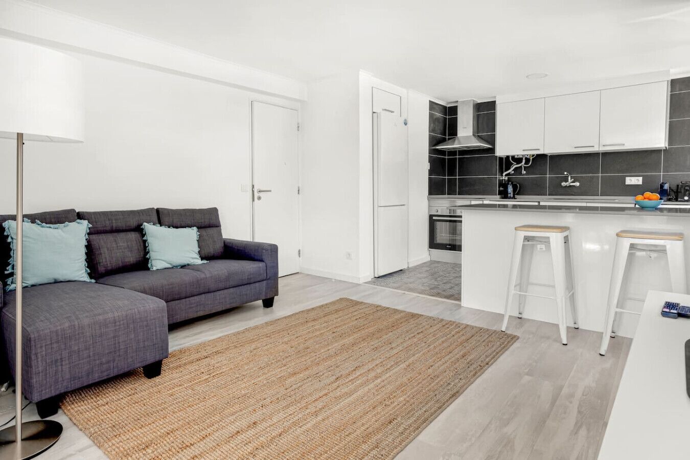 Een modern appartement met een grijze bank, een keuken met zwart-wit tegels en een kleine bar.