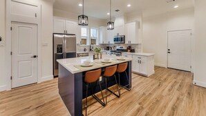 Une cuisine moderne avec des meubles blancs, un grand îlot central doté de tabourets, des appareils en acier inoxydable et un éclairage suspendu.