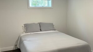 A bedroom with a single bed, a window with blinds, a ceiling light, and a dark floor. - South End Bungalow - 5 full bedrooms (Greater Sudbury)
