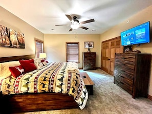Un dormitorio con cama, ventilador de techo, televisor, cómoda y ventana con persianas.