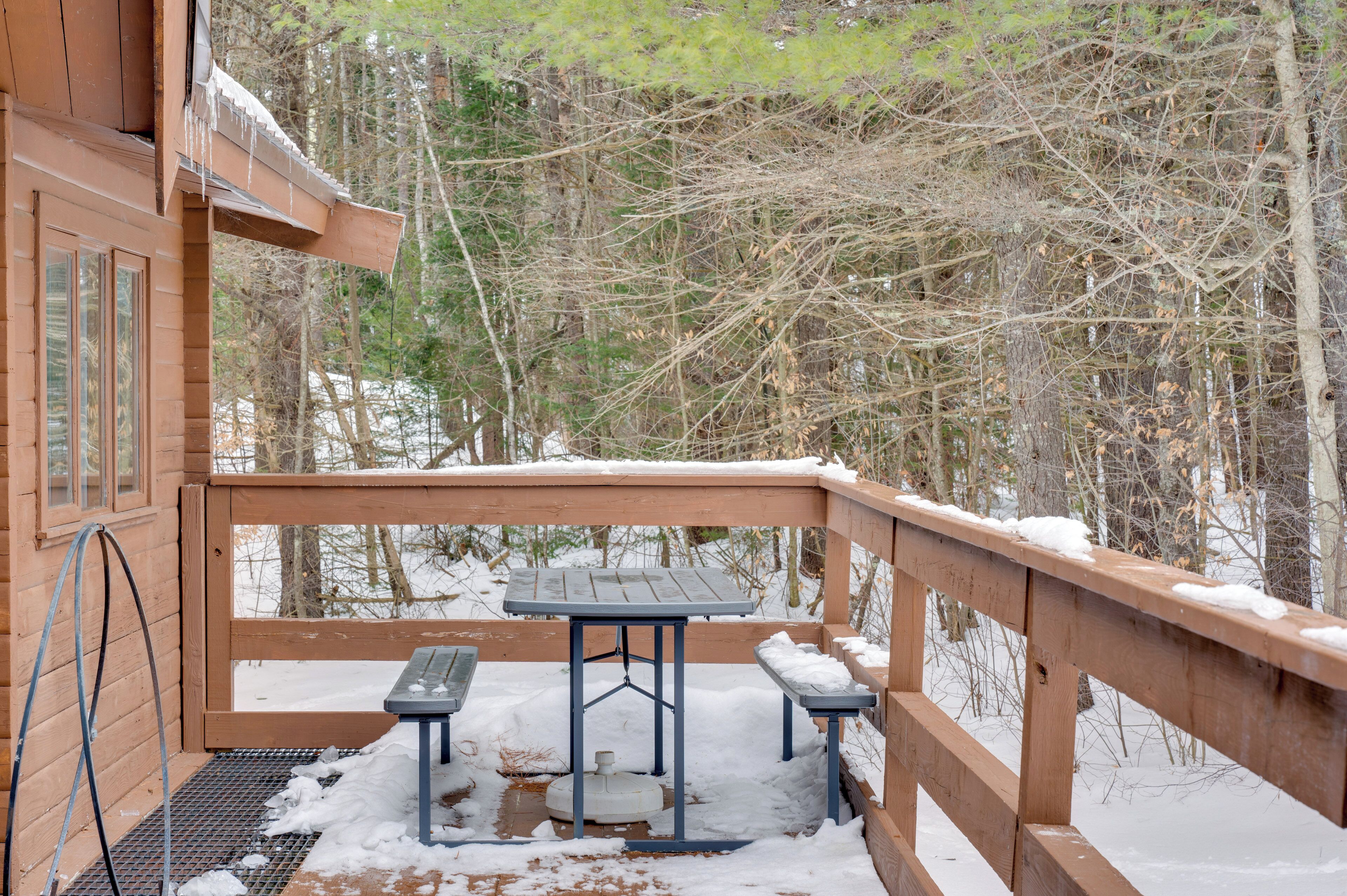 Una terraza de madera con una mesa y bancos, rodeada de nieve y árboles.