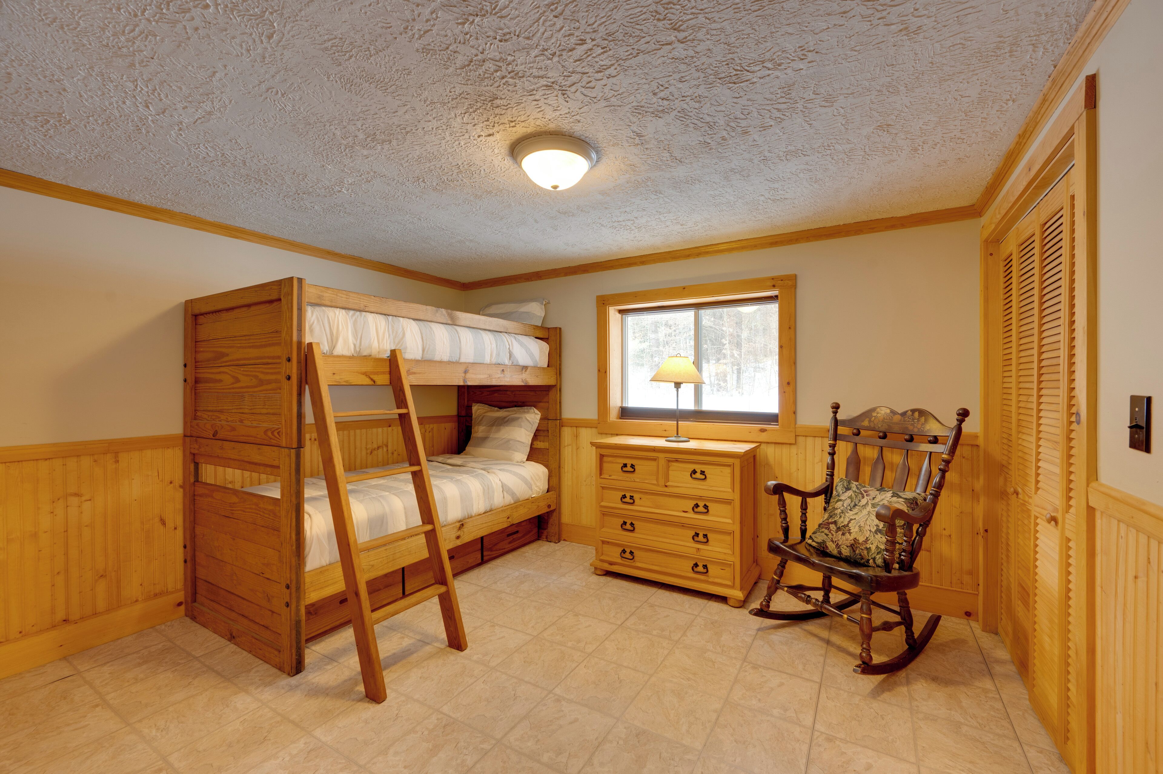 A room with a bunk bed, a dresser, a rocking chair, and a window with a lamp.