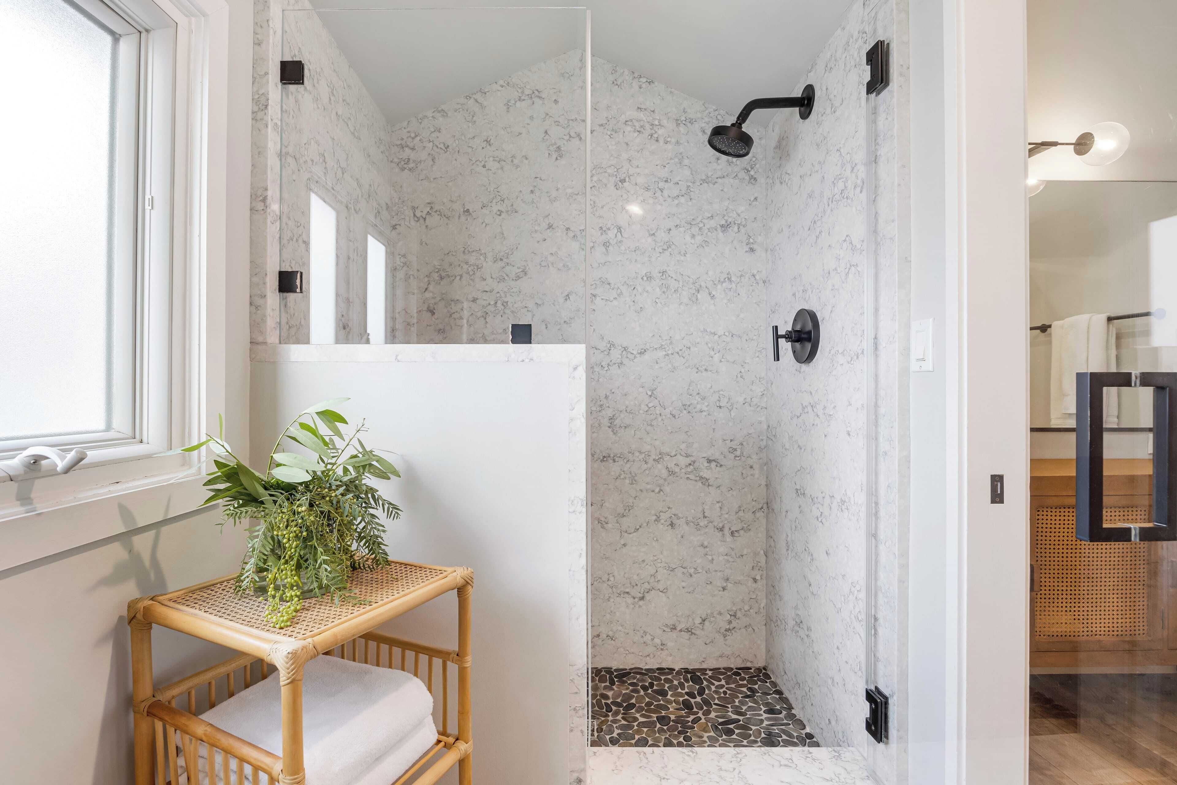 Un baño con zona de ducha que presenta una pared de mármol, una planta pequeña sobre un estante de mimbre y una ventana.
