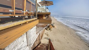 A wooden deck with a view of the beach, a wicker basket, and a beach umbrella. - Unbeatable Beachfront Bliss w/ Ocean Views, Luxe Interiors & Patio Beach Road (Dana Point)