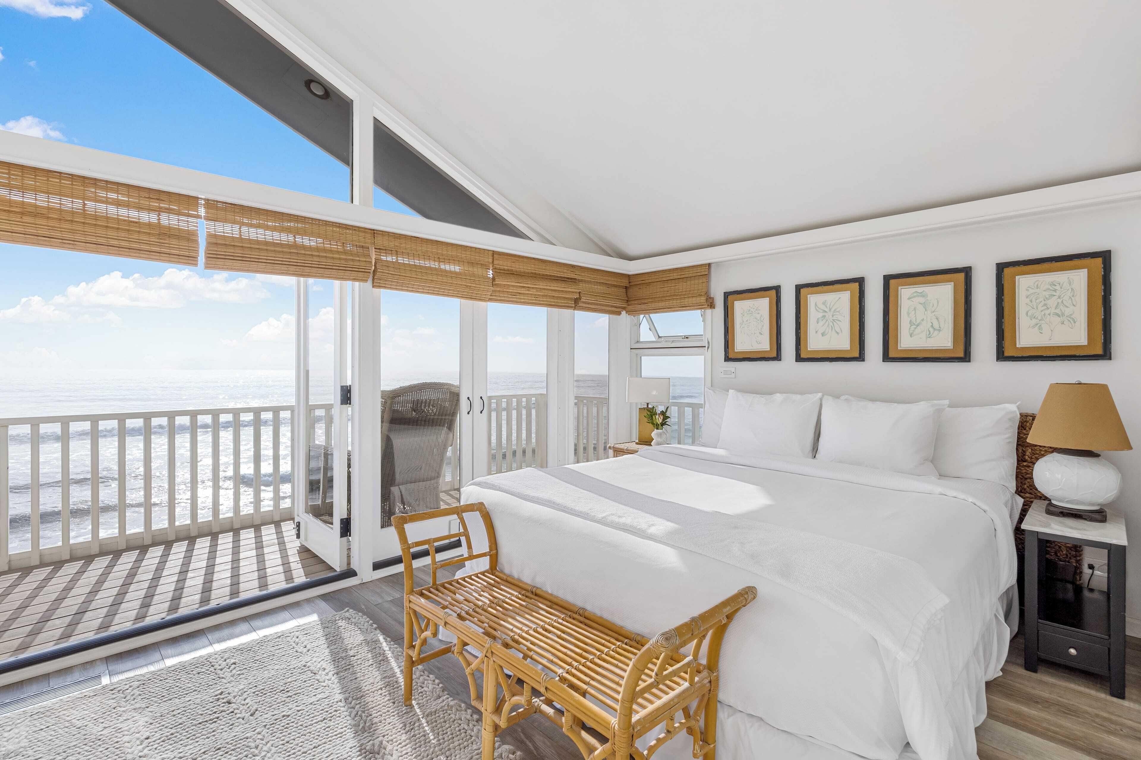 A bedroom with a large bed, a wooden bench, a nightstand, and a view of the beach.
