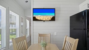 Un coin repas avec une table en bois, deux chaises, une plante en pot, un réfrigérateur noir et un téléviseur diffusant une scène de plage.