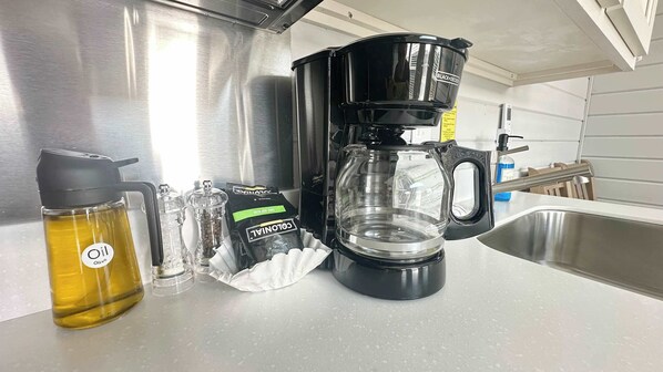 A kitchen counter with a coffee maker, a bottle of olive oil, pepper grinders, and a bag of coffee.