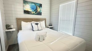 Un lit bien fait, avec du linge de lit blanc et un seul coussin décoratif, une table de chevet avec une télécommande et une œuvre d’art accrochée au mur représentant une vague.