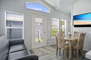 A room with a large window, a television, a dining table with chairs, and a sofa.