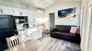 A compact kitchen with white cabinets, a black refrigerator, and a coffee maker. There's a wooden dining table and a black leather sofa with a pink pillow. A large painting of a wave is on the wall. - New Listing! Mins to Beach*2 Bdrm*Sleeps up to 6 (Bolivar Peninsula)