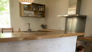 Una cocina con salpicadero de ladrillo blanco, encimera de madera, campana extractora de acero inoxidable y un taburete de madera.