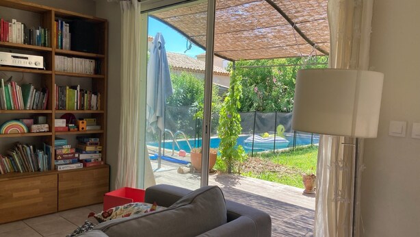 Una sala de estar con un sofá gris, una estantería llena de libros y juguetes, y vistas a una piscina y jardín a través de un ventanal.