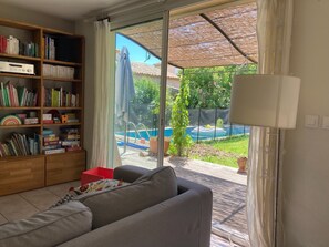 Un salon avec un canapé gris, une bibliothèque remplie de livres et de jouets, et une vue sur une piscine et un jardin, visible à travers une porte vitrée.
