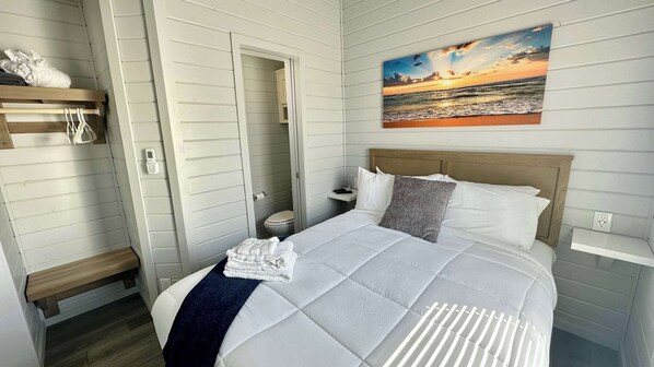 A neatly arranged hotel room with a bed, a bedside table, a nightstand, a painting of a sunset over the ocean, and a wooden bench with towels.