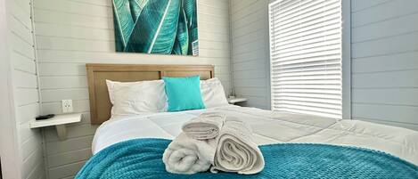 A neatly made bed with a teal blanket and rolled towels, white pillows, and a wooden headboard.