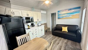 A compact kitchen with white cabinets, a black refrigerator, and a microwave. There's a wooden dining table with chairs, a black leather sofa with a yellow pillow, and a painting of a beach scene on the wall. - Pet Friendly*Near Beach & Margaritaville (Bolivar Peninsula)