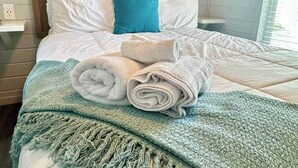 A neatly made bed with rolled towels, a blue and white throw blanket, and a painting of a wave on the wall. - Pet Friendly*Near Beach & Margaritaville (Bolivar Peninsula)