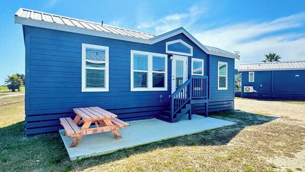 Una casa azul de una sola planta, pequeña, con un porche, un banco de madera y una escalera que lleva a la entrada.