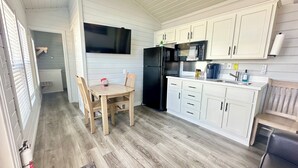 Una cocina compacta con armarios blancos, un refrigerador negro, una mesa de comedor de madera con sillas y una ventana pequeña.