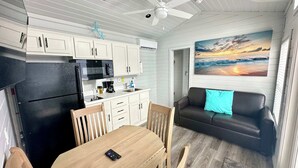 A compact kitchen with white cabinets, a black refrigerator, and a microwave. There's a dining table with chairs and a black leather sofa with a blue pillow. A painting of a sunset is on the wall.