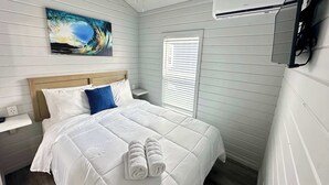 A neatly made bed with white bedding and a blue pillow, a painting of a wave on the wall, and a window with blinds.