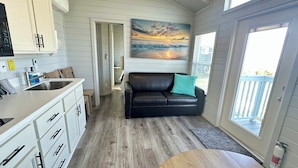 A compact kitchen with a sink, cabinets, and a countertop. There is a black leather sofa with a teal pillow and a painting of a sunset above it. A small table is in the foreground.