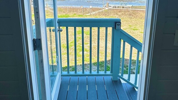 A view from a room's door to a balcony with a railing, a fire extinguisher, and a clear sky.