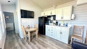 Cocina compacta con armarios blancos, un refrigerador negro y un microondas. Hay una mesa de comedor con sillas y un ventanal pequeño.