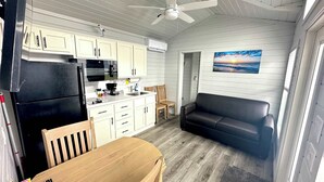 Una cocina compacta con armarios blancos, un refrigerador negro y un microondas. Hay una mesa de comedor de madera y un sofá negro en una sala de estar con un cuadro de un atardecer.