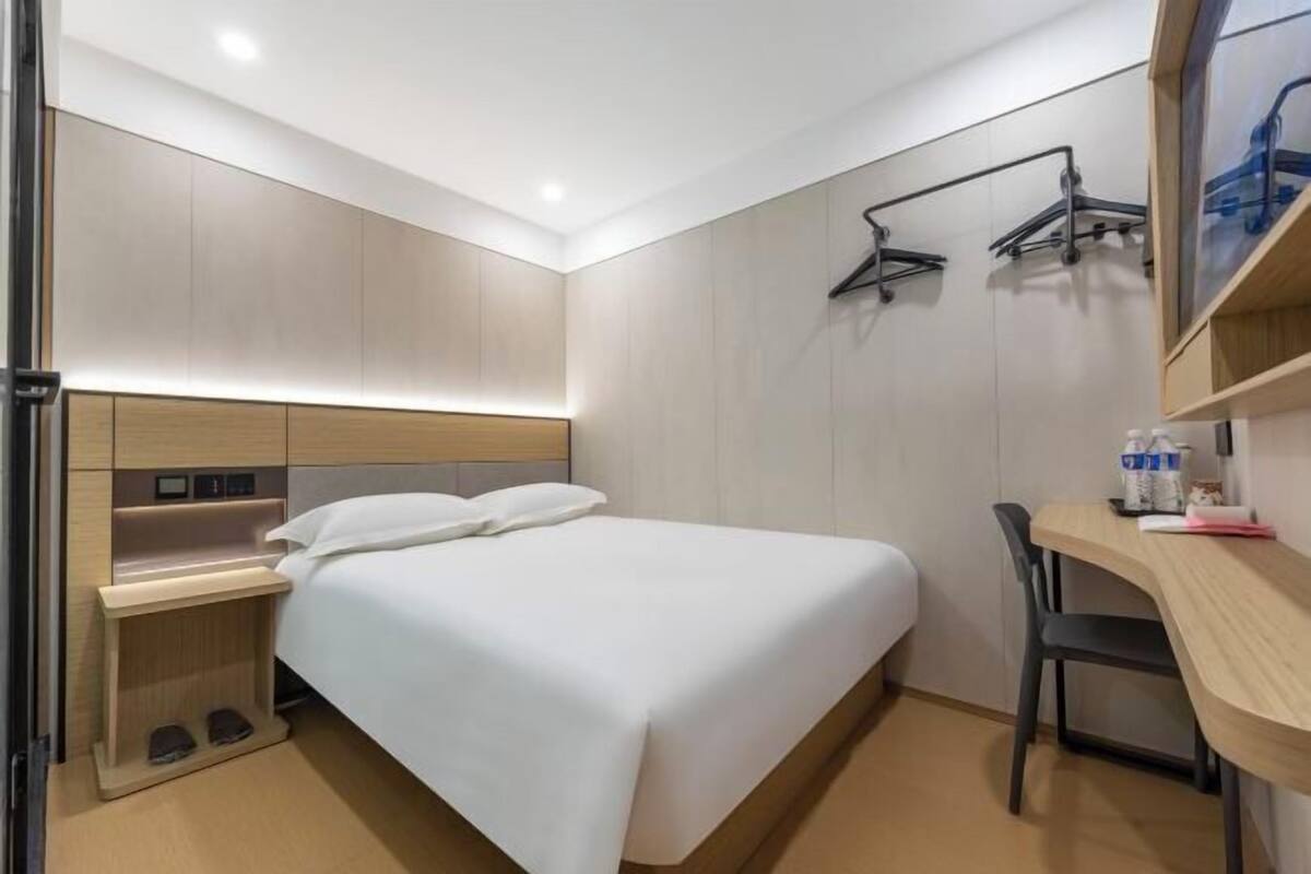 A modern hotel room with a bed, bedside table, chair, and a desk with a chair.