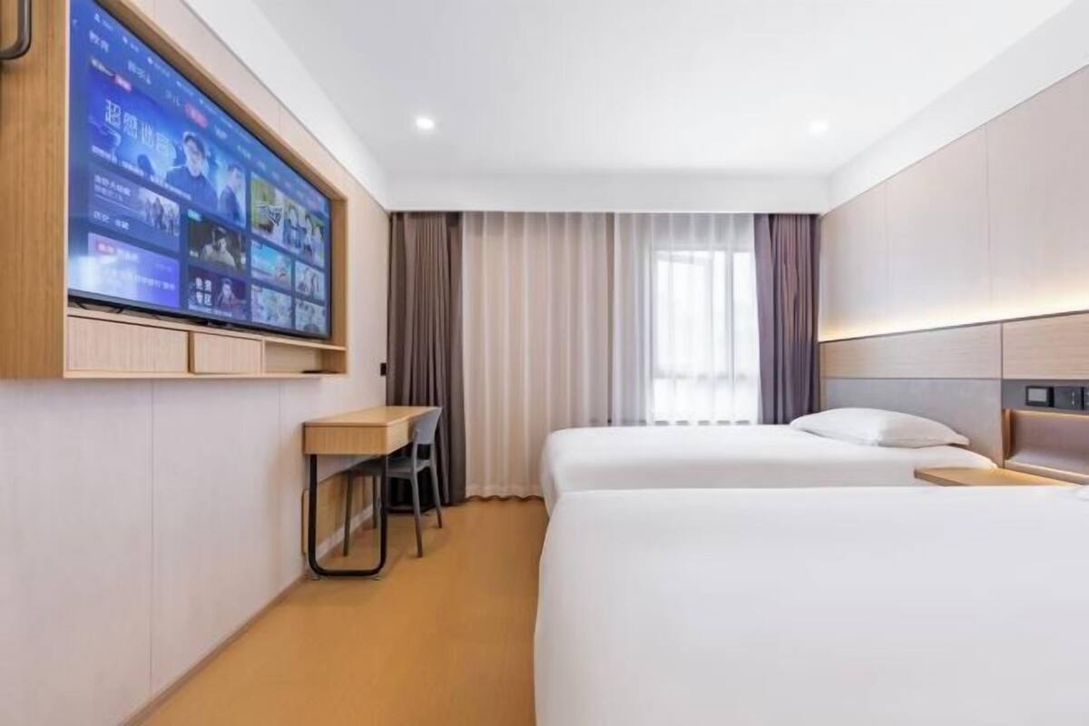 A hotel room with a large flat-screen TV mounted on the wall, a desk with a chair, two beds, and a window with curtains.