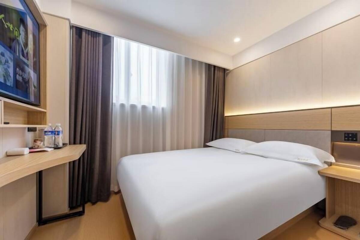 A modern hotel room with a large bed, a bedside table, a built-in shelf with toiletries, and a large window with curtains.