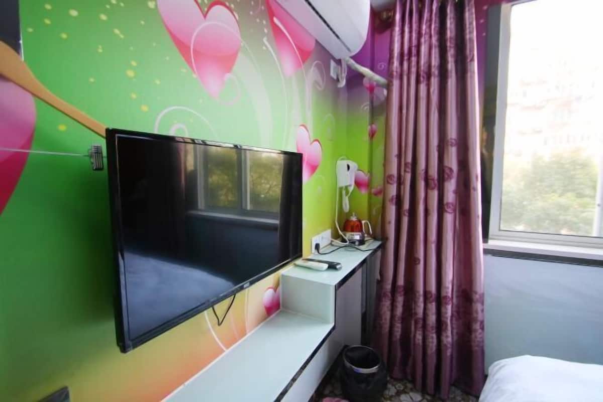 Une pièce avec un téléviseur fixé au mur, un bureau avec une lampe et une fenêtre avec des rideaux.