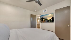 A hotel room with a bed, a ceiling fan, a painting of a sunset, and a view into the kitchen area. - Oak's Beautiful Contemporary Suite #06 (Lawton)