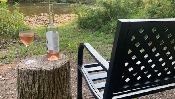 Un banc avec une bouteille de vin et un verre de vin, posés sur une souche d’arbre au bord d’une rivière.