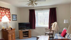 Un salon avec un ventilateur de plafond, un téléviseur, un meuble en bois, un canapé à carreaux et un fauteuil rouge.