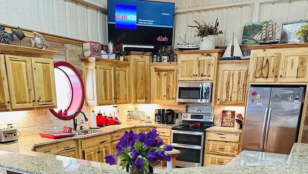 A kitchen with wooden cabinets, a granite countertop, a built-in island with bar stools, a stainless steel refrigerator, and a flat-screen TV displaying a CD.