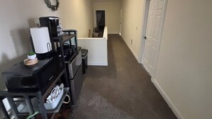 Un pasillo estrecho con una zona de cocina a la izquierda, que incluye una cafetera y un cubo de basura. Se ve una puerta a la derecha.