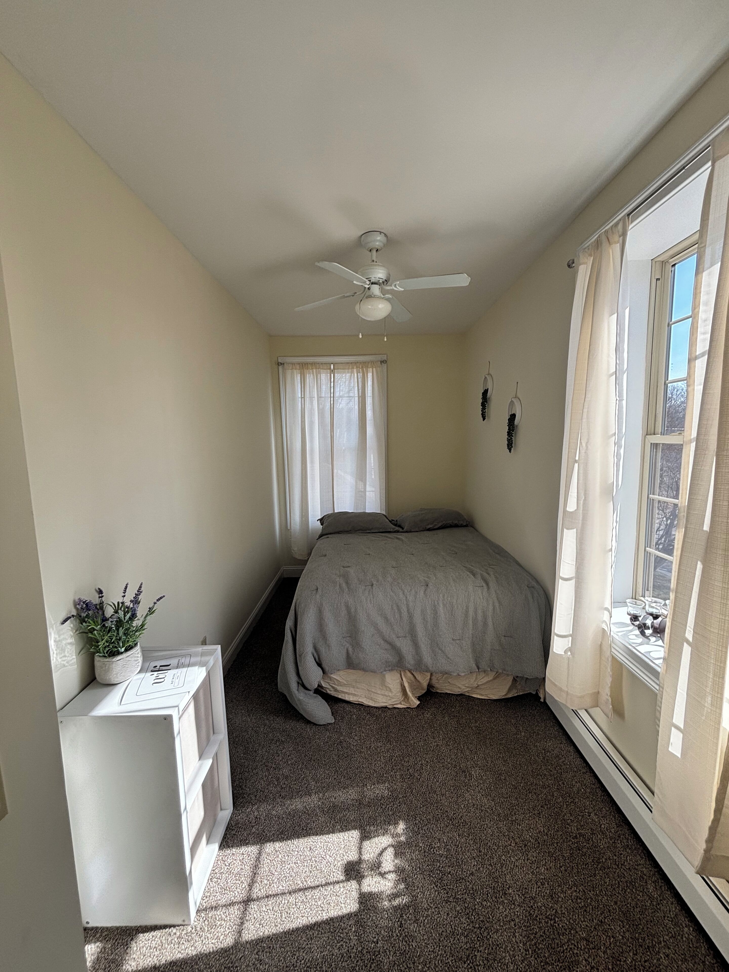 Un dormitorio con cama, ventilador de techo, ventana con cortinas y una pequeña mesa con una planta.