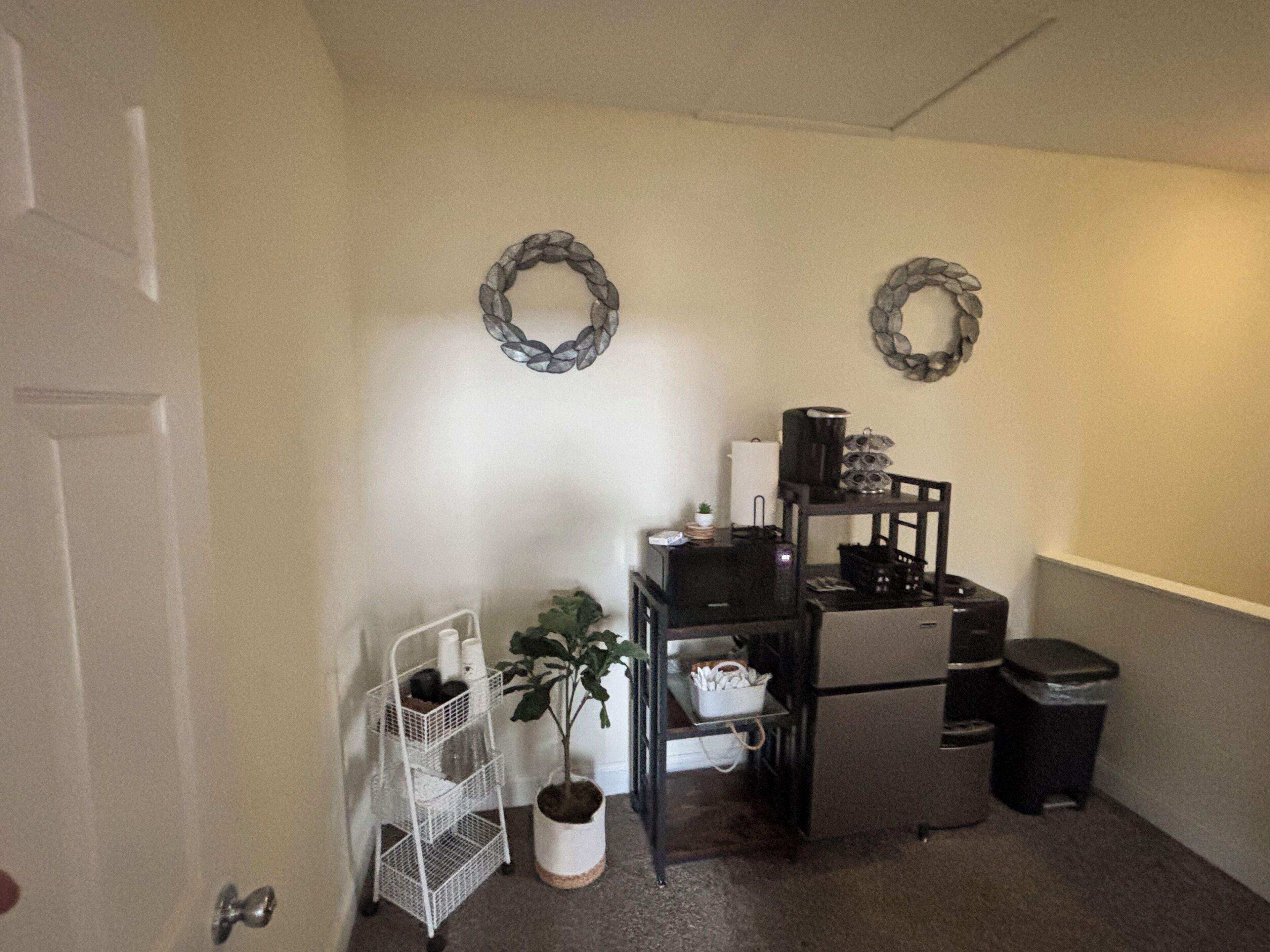 A compact kitchen with a refrigerator, stove, and a coffee maker on a small table. There is a white wire shelf with items and a potted plant. Two decorative wreaths adorn the walls.