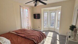 Un dormitorio con cama, ventilador de techo, televisor montado en la pared y ventana con vista a árboles.