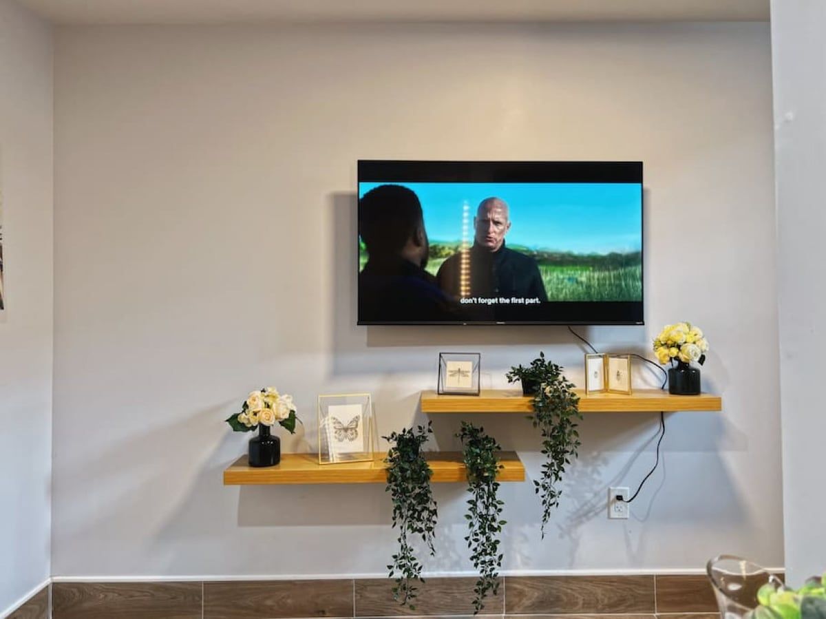 Een televisie aan een muur bevestigd, met daaronder een plank waarop ingekaderde foto’s en bloemen staan.
