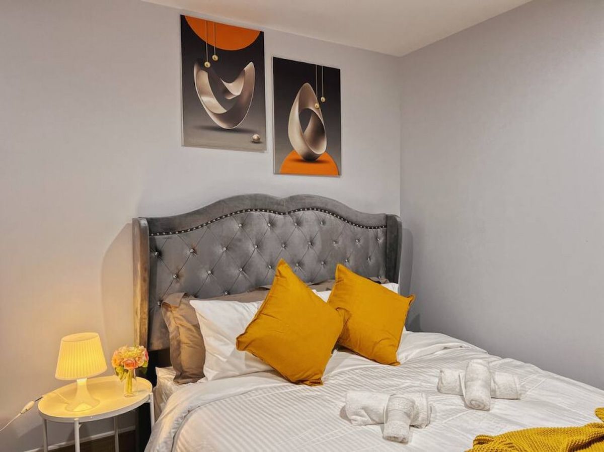 A hotel room with a tufted headboard, a bed with white linens and two mustard pillows, a bedside table with a lamp and flowers, and abstract artwork on the wall.