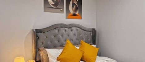 A hotel room with a tufted headboard, a bed with white linens and two mustard pillows, a bedside table with a lamp and flowers, and abstract artwork on the wall.