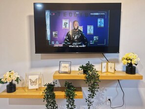 Un televisor montado en la pared mostrando a una persona con el rostro desenfocado, rodeada de fotografías enmarcadas y un estante con objetos decorativos.