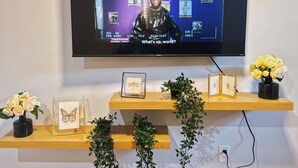 A television mounted on a wall displaying a sports event with a person in a black outfit. Below the TV, there are wooden shelves with framed pictures and a vase of flowers. - Ramblei near Chinatown & CenterCity: 1 Bedroom - Unit A (Philadelphia)